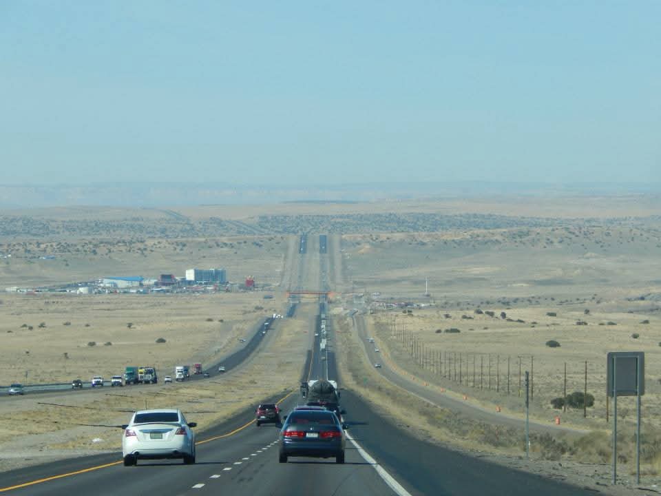 A long, straight highway stretching into the distance with vehicles traveling on both sides, surrounded by open desert land and a clear blue sky.