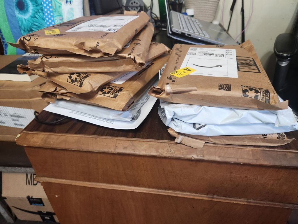 A stack of assorted packages in brown paper and plastic on a wooden desk, with a laptop in the background.