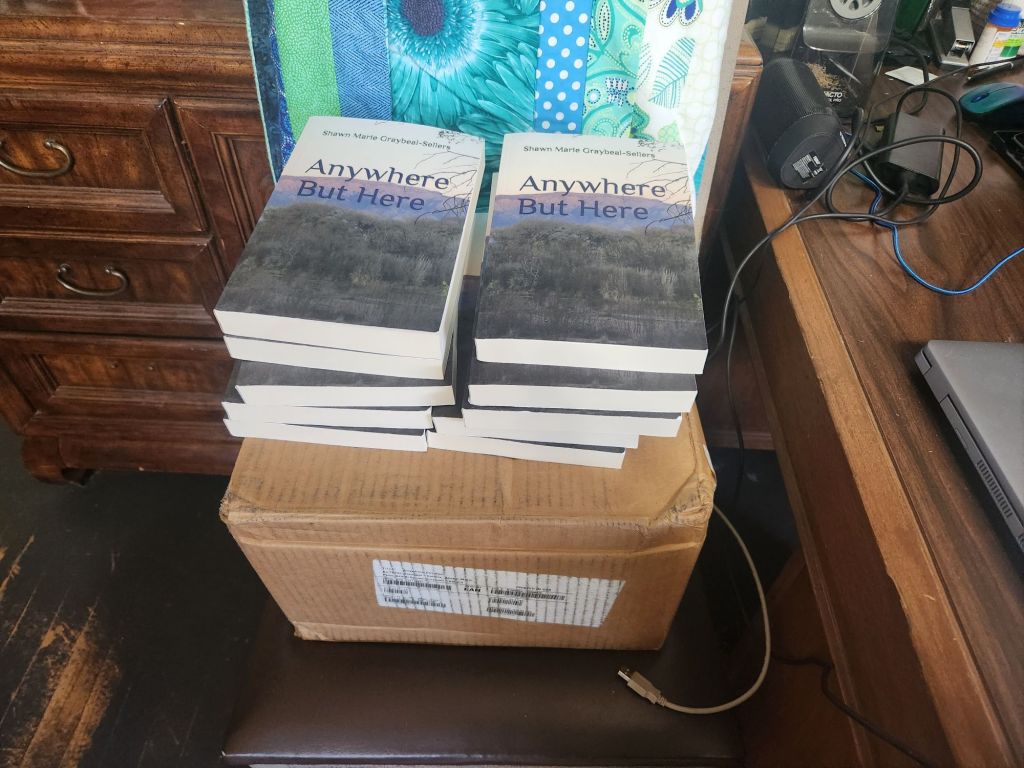 A stack of books titled 'Anywhere But Here' by Shawn Marie Graybeal-Sellers placed on top of a cardboard box, with a wooden desk and various items in the background.