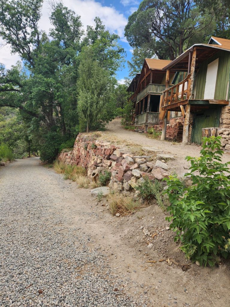 A gravel path leading towards rustic buildings nestled among trees and greenery.
