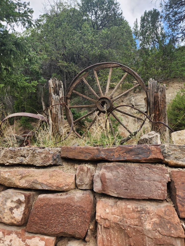 An old wooden wagon wheel resting against a weathered stone wall, surrounded by greenery and trees.