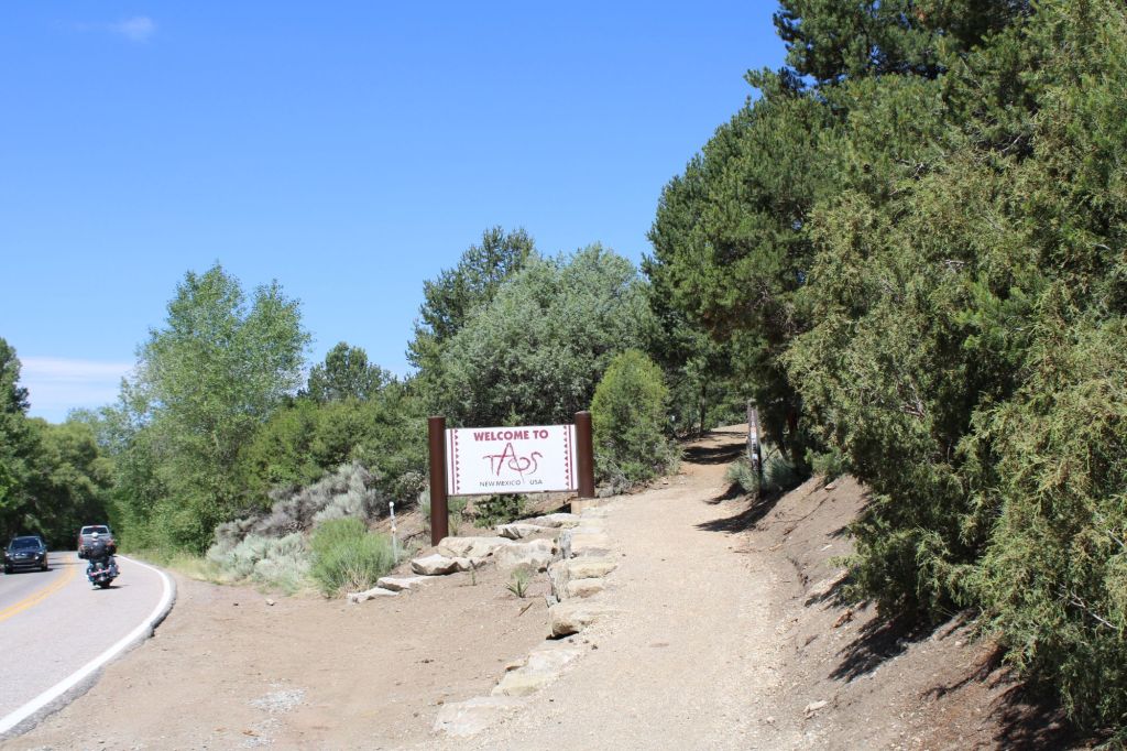 Welcome sign for Taos, New Mexico, alongside a road surrounded by greenery and a dirt path.