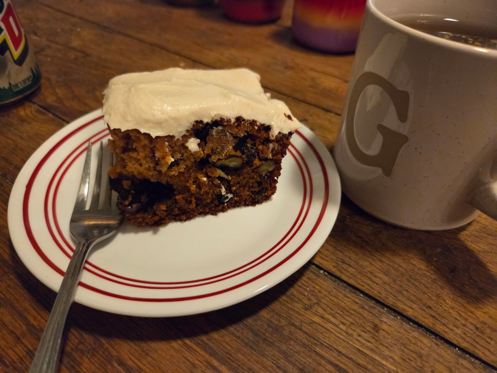 A slice of cake with frosting on a white plate next to a coffee mug, set on a wooden table.