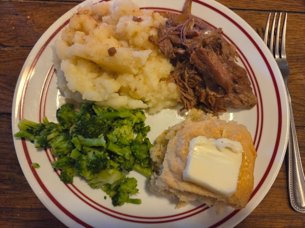 A plate with mashed potatoes, pulled meat, steamed broccoli, and a buttered biscuit, set on a wooden table.